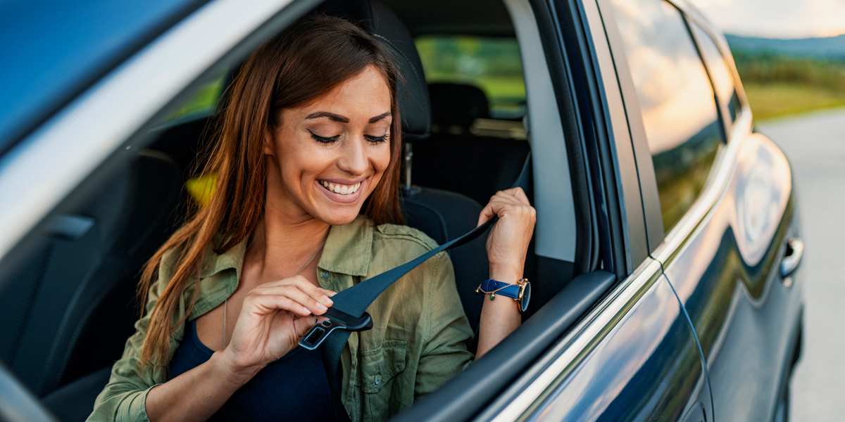 Eine Frau sitzt im Auto auf dem Fahrersitz und schnallt sich gut gelaunt an. 