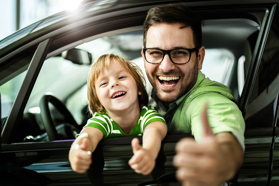 Mann sitz mit seinem Sohn im Auto und guckt aus dem heruntergelassenen Fenster auf der Fahrerseite. Beide halten die Daumen hoch und sind glücklich.