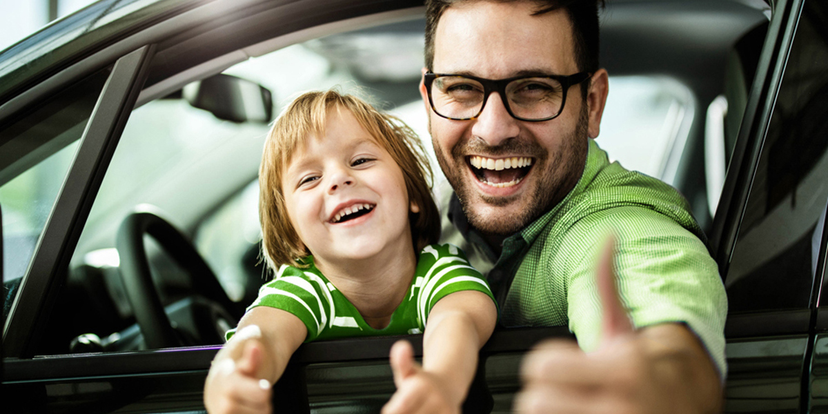 Mann sitz mit seinem Sohn im Auto und guckt aus dem heruntergelassenen Fenster auf der Fahrerseite. Beide halten die Daumen hoch und sind glücklich.